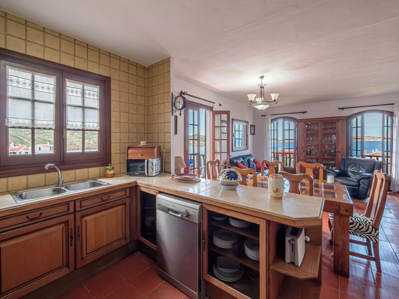 A kitchen with wooden cabinets, a dishwasher, a dining table with chairs, and a view of the sea through large windows.