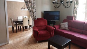 Una sala de estar con piso de madera, un sillón rojo, un sofá rojo, un televisor sobre un mueble, una araña y un comedor con mesa y sillas.