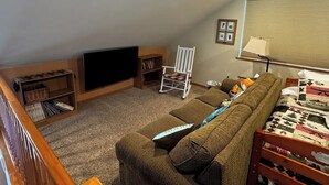 A cozy living room with a sectional sofa, a white rocking chair, a flat-screen TV, and a bookshelf.