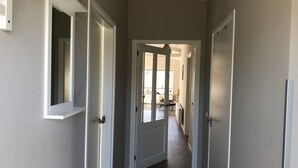 Un couloir avec des portes blanches, un luminaire au plafond et une vue sur une autre pièce où l’on aperçoit une chaise et une table.