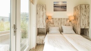 A bedroom with two beds, wooden headboard panels, wall-mounted lamps, and a view of greenery through a glass door. - Comfort Lodge in Boomhiemke (Hollum-Ameland)