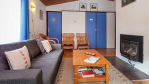 Een gezellige woonkamer met een bank, twee rieten fauteuils, een salontafel met boeken, een open haard en een blauwe deur.