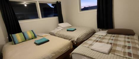 A room with three single beds, each with a different patterned bedspread. There are windows with black curtains, and the room has a wooden floor.