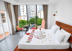 A hotel room with a large bed, a balcony with a table and chairs, and a view of buildings and greenery. - Bliss & Beats Hostel (Phnom Penh)