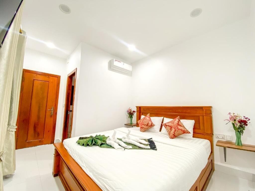 Quarto de hotel com cama de madeira, roupa de cama branca, mesa de cabeceira com um vaso de flores e porta de madeira.