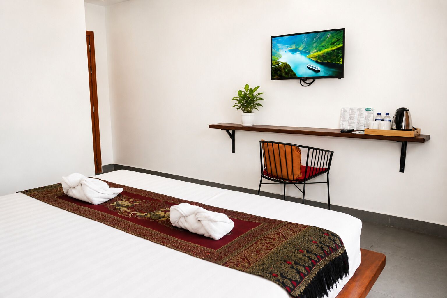 Habitación de hotel con cama, televisión montada en la pared, escritorio pequeño con silla, planta y estante con artículos de aseo.