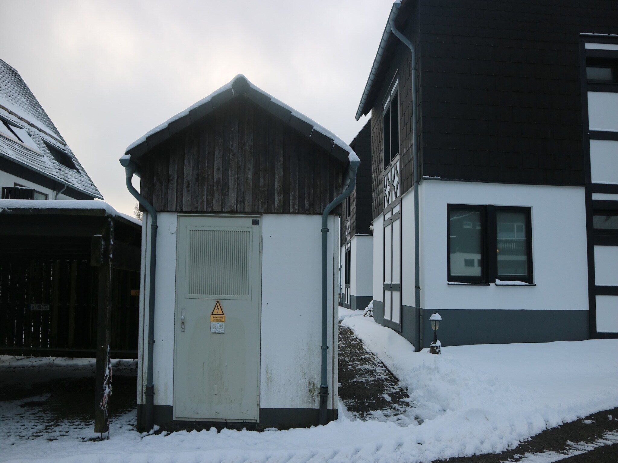 Un piccolo edificio di servizio con un cartello di avvertimento, vicino a una casa con un tetto ripido e una finestra.