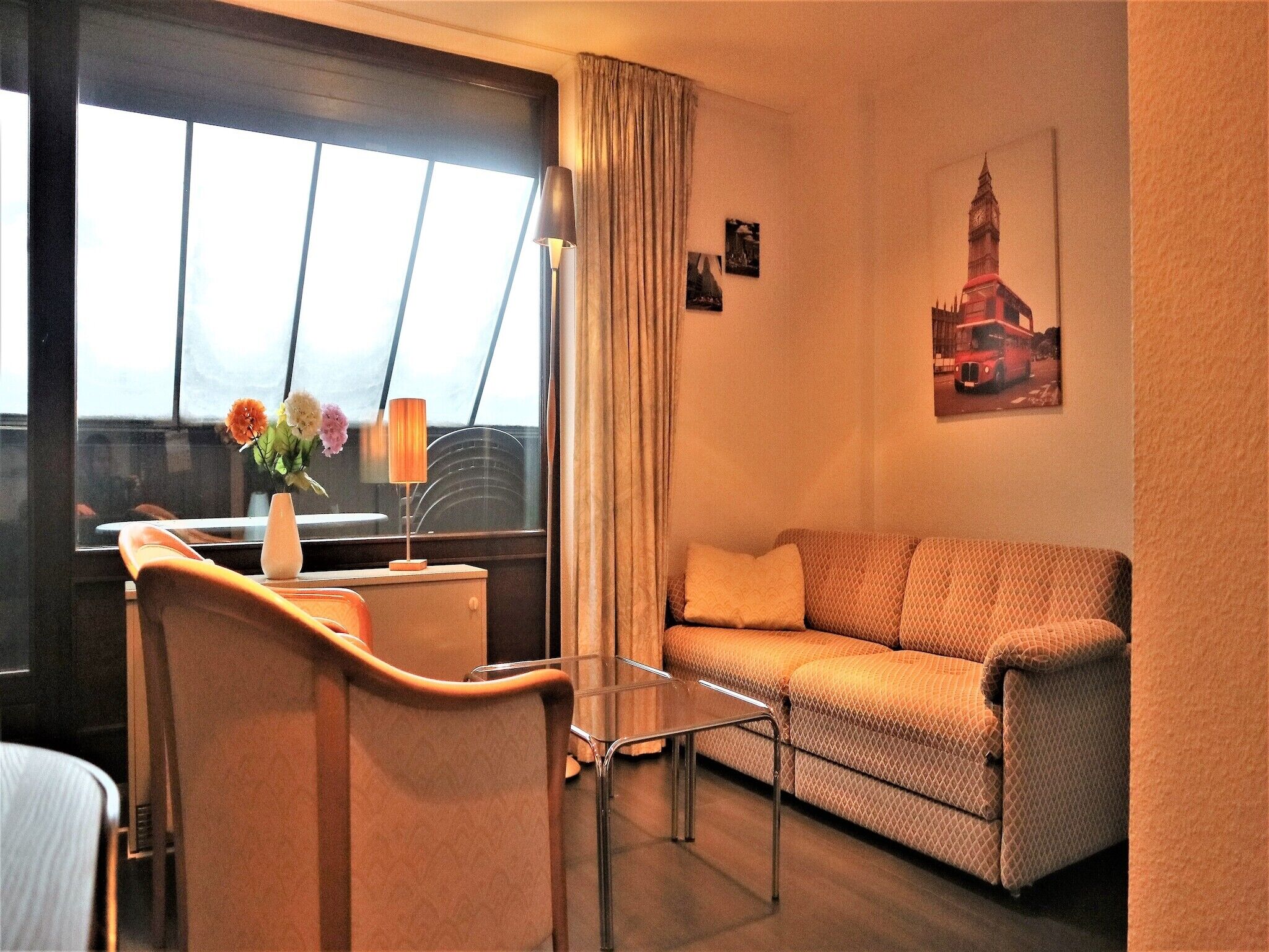 A hotel room with a sofa, a small table with a vase of flowers, a lamp, and a window with curtains.