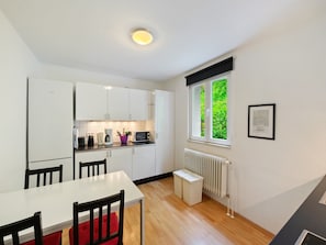 A compact kitchen with white cabinets, a dining table with chairs, a window with a view of greenery, and a radiator.