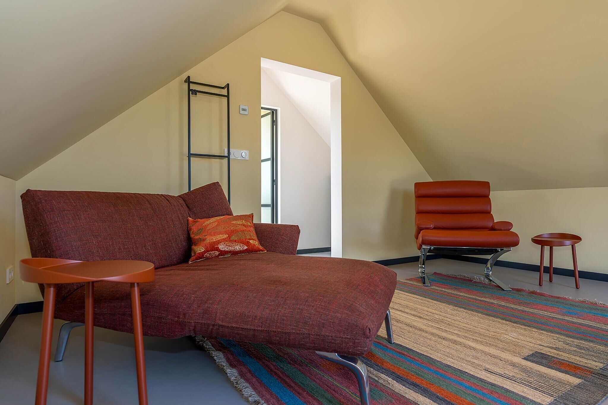 Ein gemütliches Schlafzimmer im Loft-Stil mit einem roten Schlafsofa, einem roten Sessel und einem kleinen Hocker. Auf dem Boden liegt ein farbenfroher Streifen Teppich.