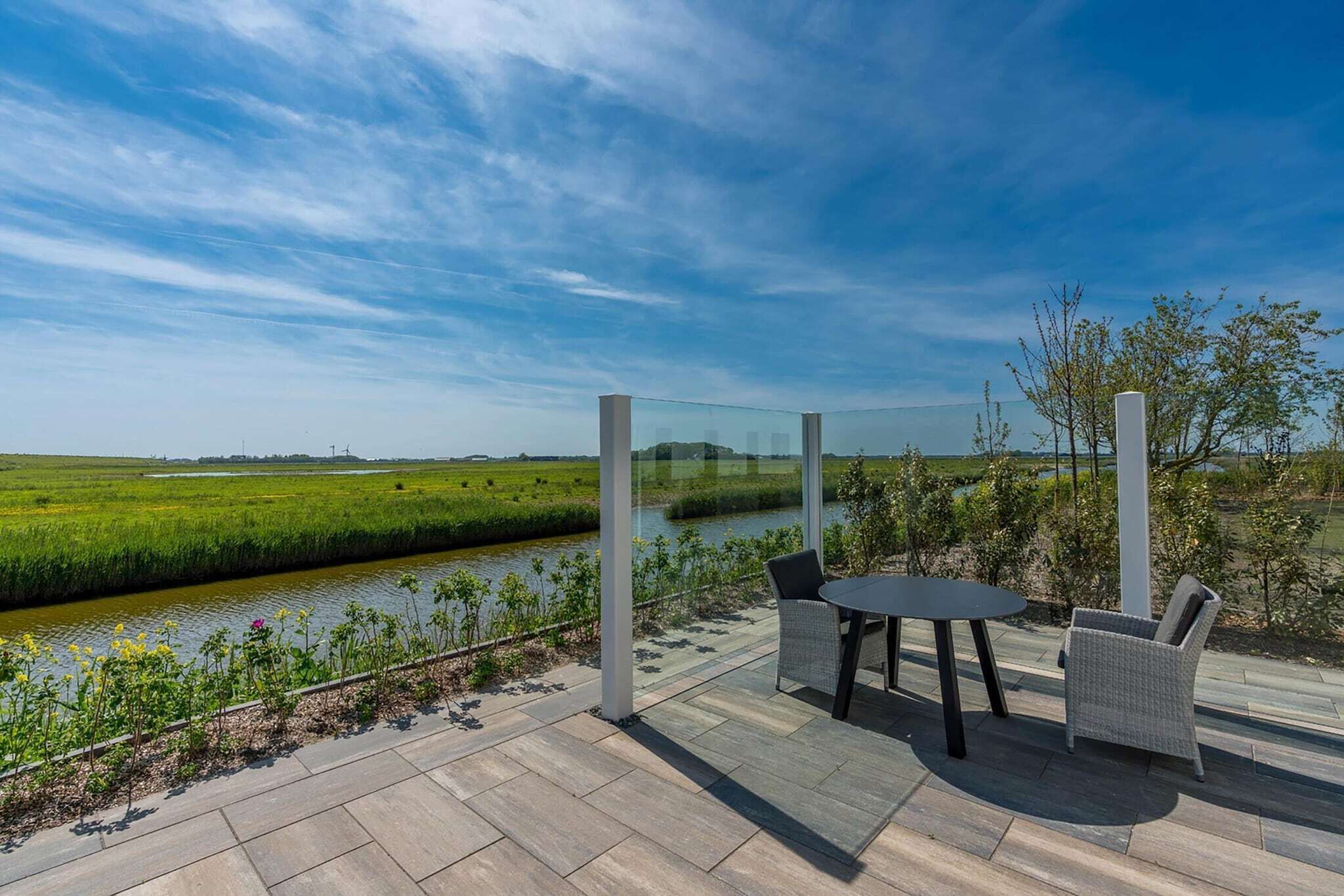 A terrace with a glass enclosure, a table, and two chairs overlooking a river and green fields.