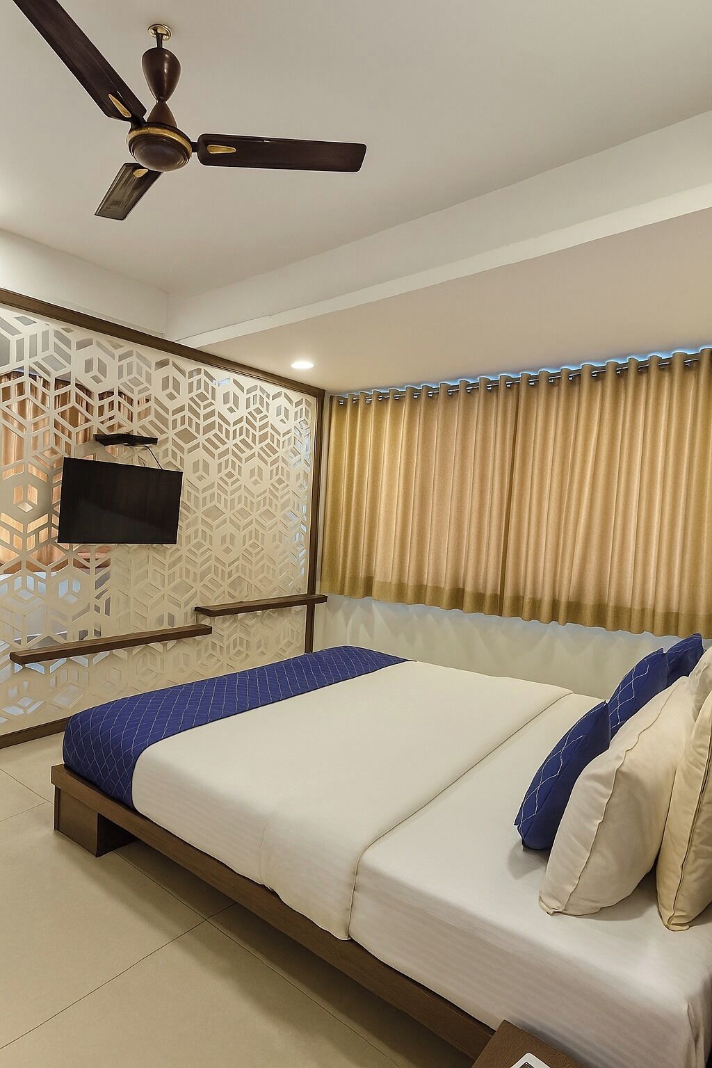 A hotel room with a bed, a ceiling fan, a TV mounted on a decorative wall, and a window with curtains.