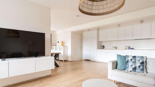 A modern living room with a flat-screen TV, a white sofa with patterned cushions, and a round coffee table.