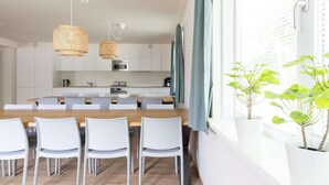 Cozinha moderna com armários brancos, uma ilha de madeira e cadeiras brancas. Há duas luminárias de palha pendentes e uma janela com cortinas.