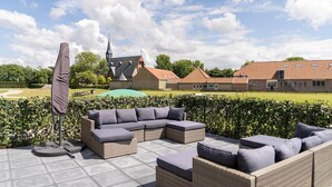 Um terraço ao ar livre com móveis de vime e um guarda-sol, um campo de golfe ao fundo e um edifício com telhado de telhas vermelhas.