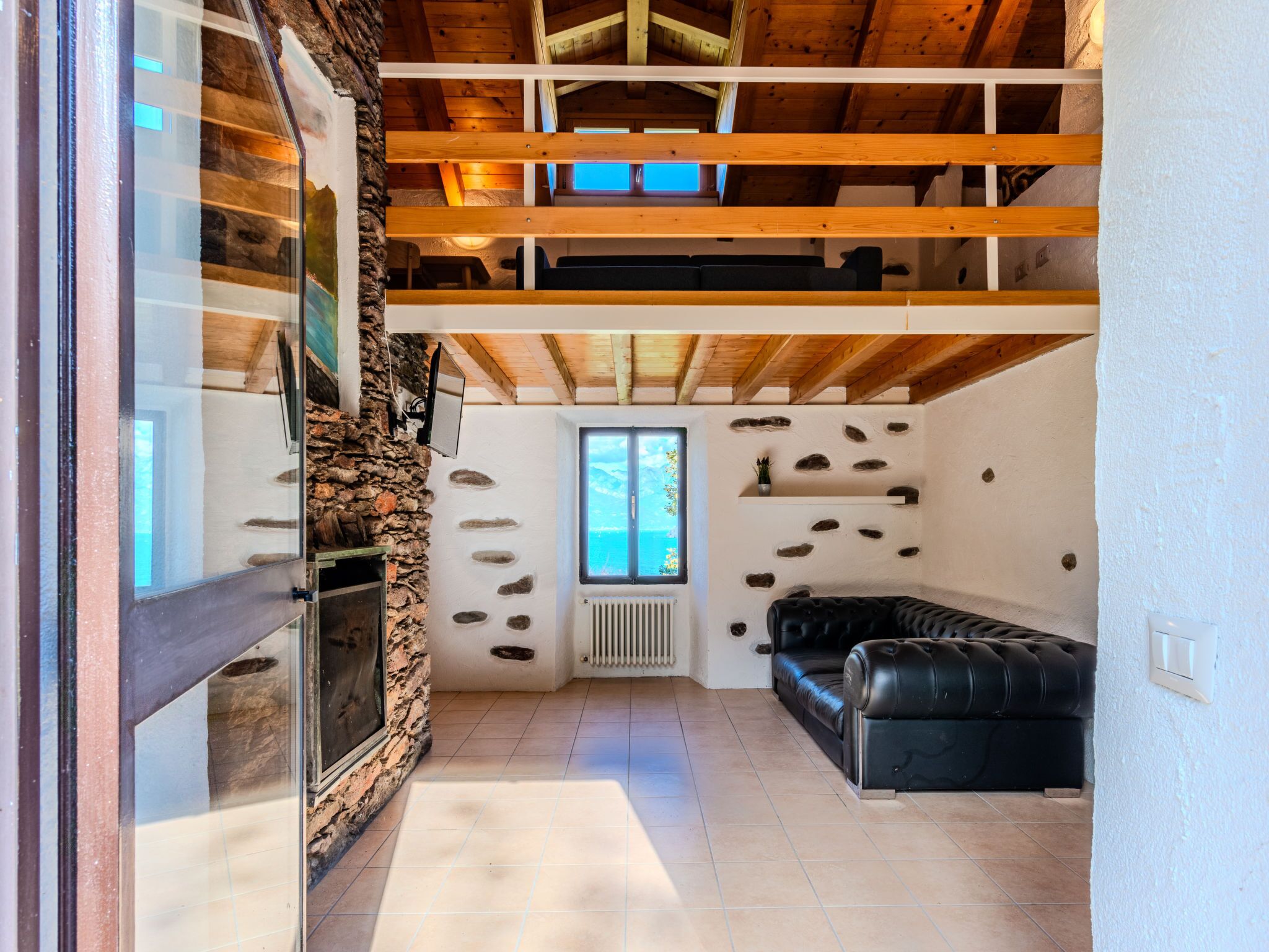 Una habitación amplia con pared de piedra, sofá de cuero negro, techo de madera y un ventanal grande.