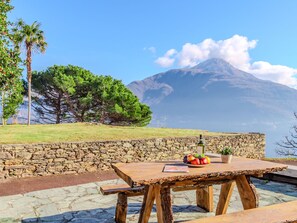 Une table en bois rustique, sur laquelle sont posés un panier de fruits et une bouteille, le tout placé devant un mur de pierre offrant une vue sur la montagne.