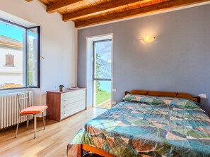 Une chambre avec un plafond en bois, un lit recouvert d’une courtepointe, une chaise, une commode et une fenêtre donnant sur un bâtiment et de la verdure.