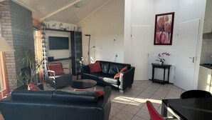Una sala de estar con un sofá de cuero negro, un sillón negro y una mesa de centro de vidrio. Se observa una mesita con una planta en maceta y un cuadro en la pared.