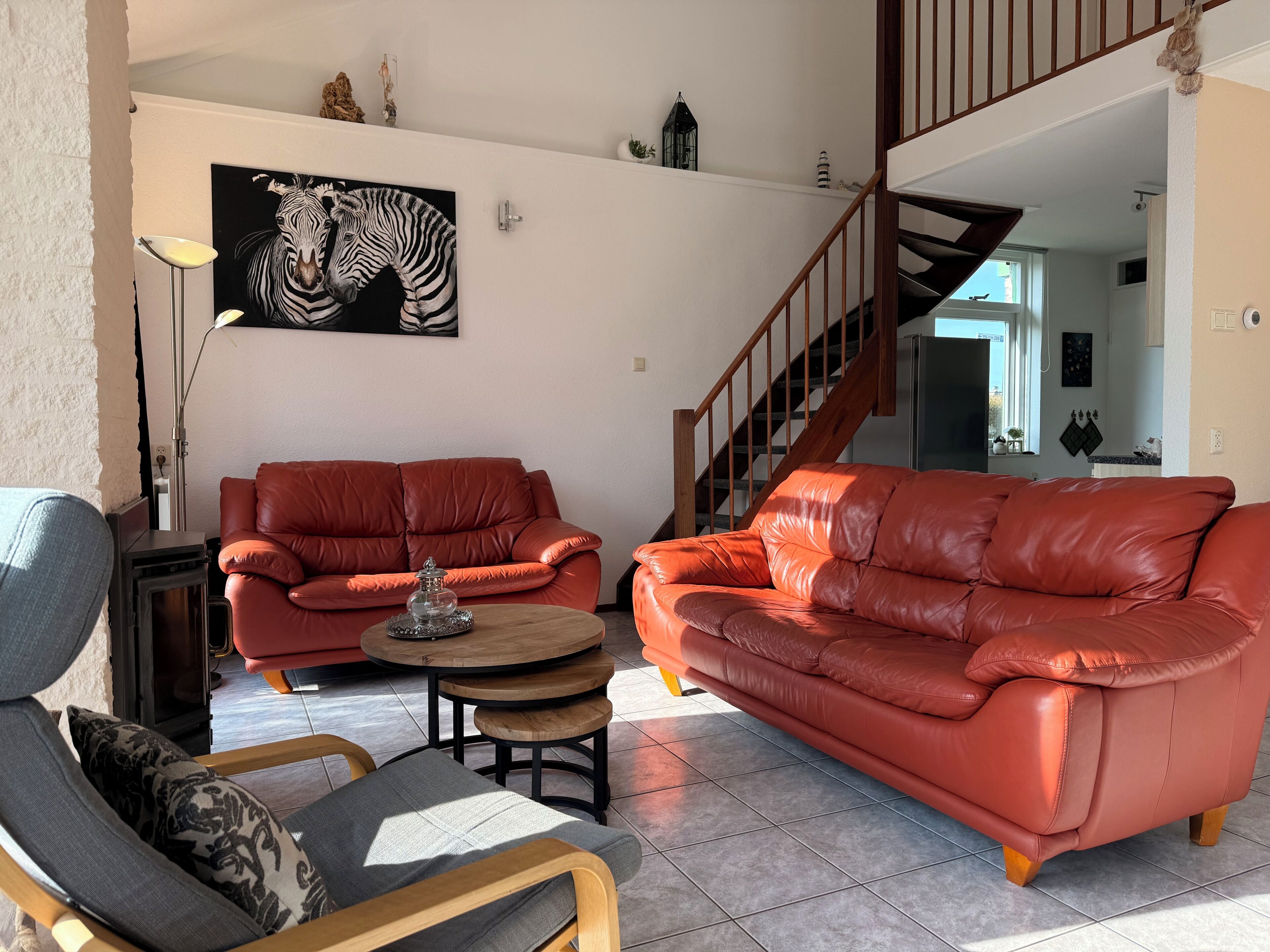 Een woonkamer met twee rode leren banken, een houten salontafel, een grijze fauteuil en een schilderij van een zebra aan de muur.