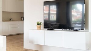A modern living room with a flat-screen TV mounted on a white cabinet, a small potted plant, and a kitchen in the background. - Group Apartment Ameland near Nes (Nes)