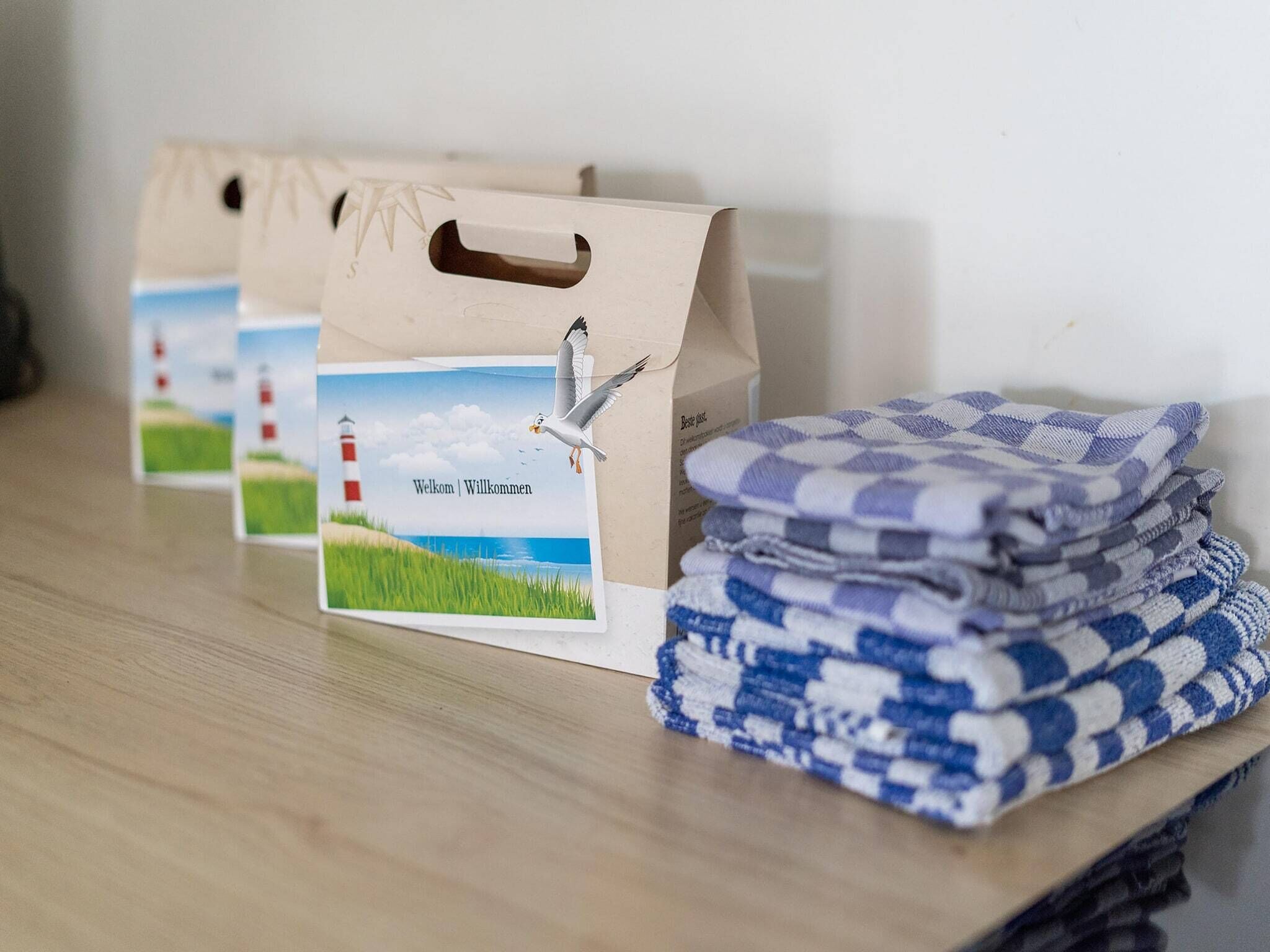 A stack of folded towels with a checkered pattern and a paper bag with a lighthouse image and the words "Welcome" on a wooden surface.