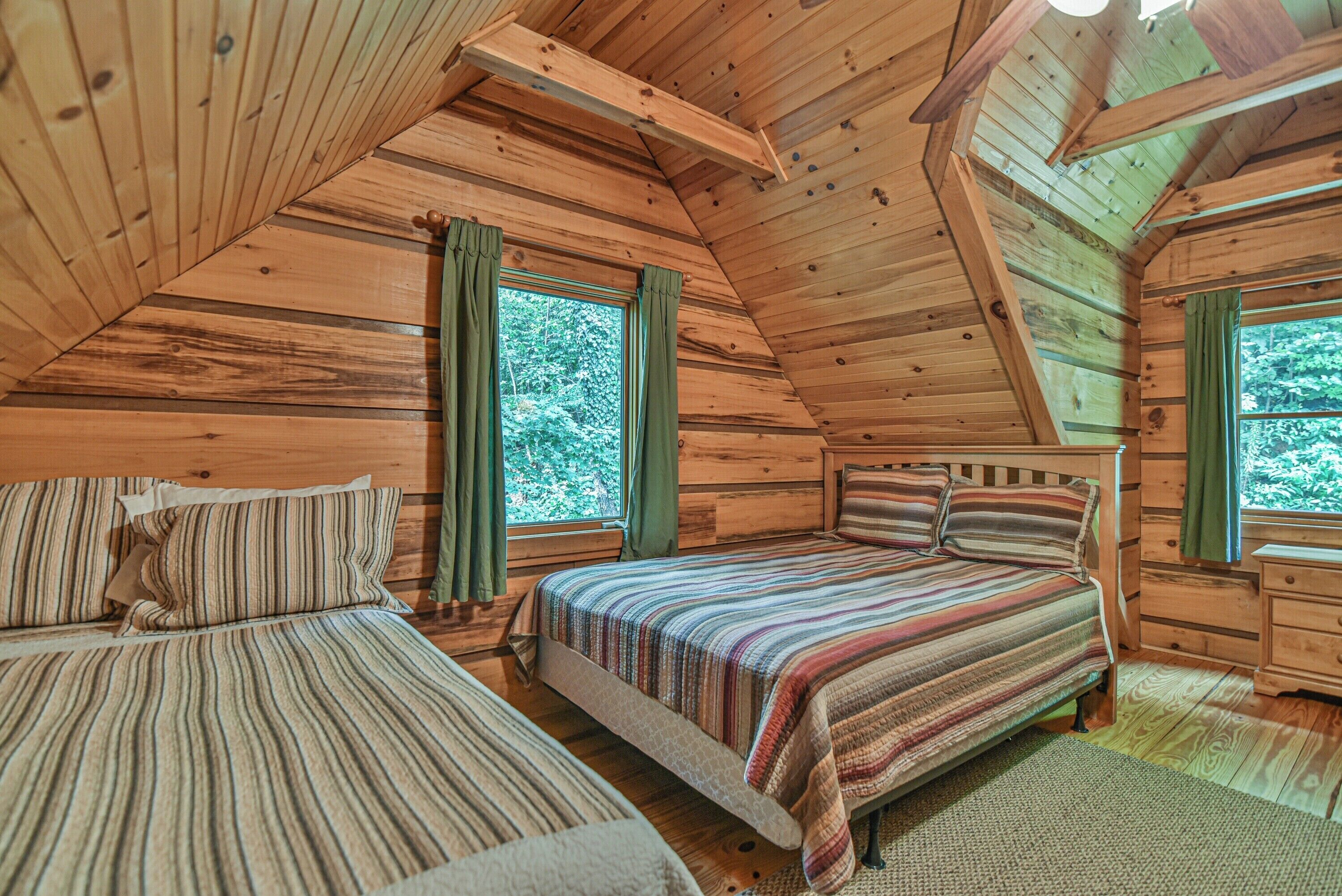 A cabin with two wooden beds, a wooden ceiling, and large windows with curtains.
