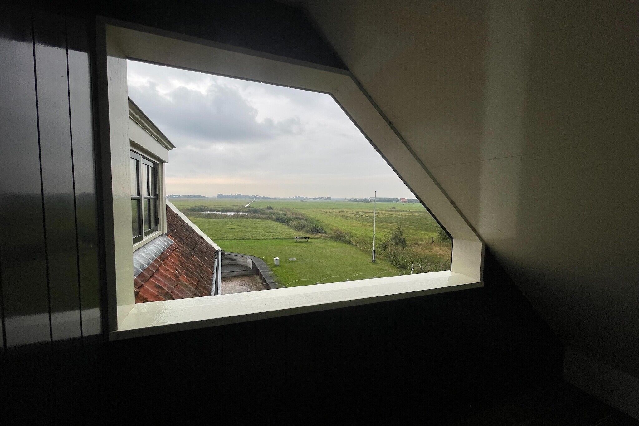 A view from a window with a sloped roof, overlooking a green field and a distant building.