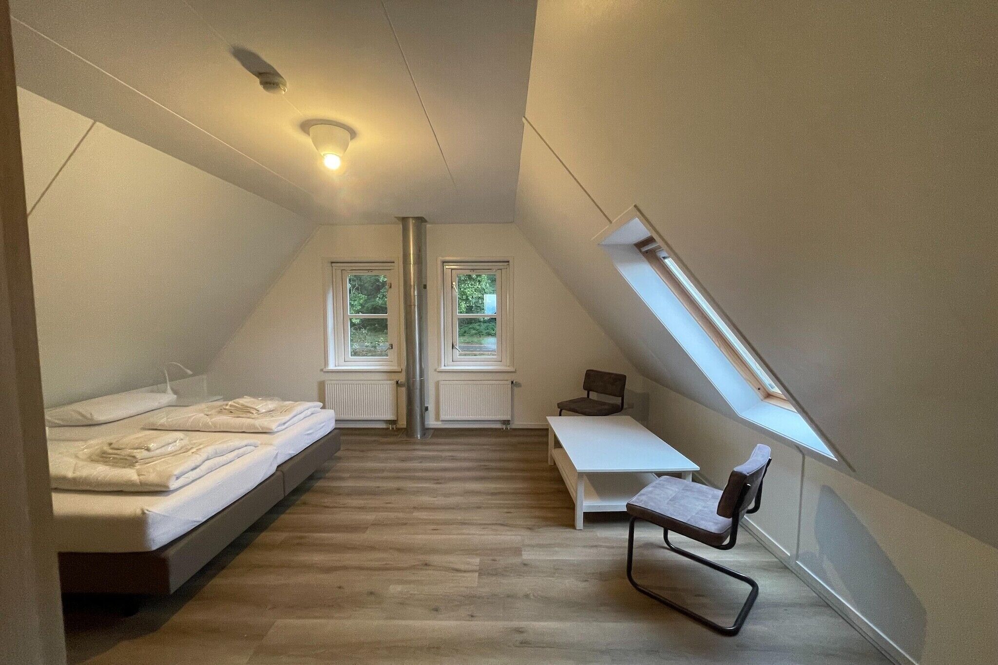 Une petite chambre moderne située sous les combles, avec un lit en mezzanine, un bureau et une chaise. Deux fenêtres laissent entrer la lumière naturelle.