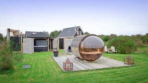 A modern house with a large wooden barrel structure in the backyard, a stone wall, and a wooden deck.