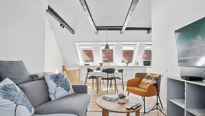 Ein modernes Wohnzimmer mit einem grauen Sofa, einem Couchtisch und einem orangefarbenen Sessel. Es gibt einen Essbereich mit Tisch und Stühlen, und im Hintergrund eine Küche.