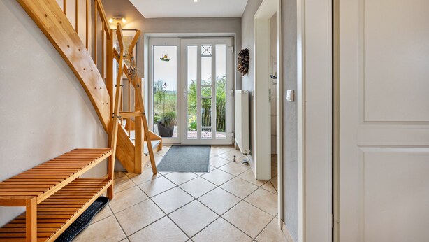 Een trap met houten treden die leidt naar een deur, een betegelde vloer en een uitzicht door glazen deuren op een buitenruimte.