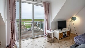 Une pièce dotée d’une porte-fenêtre coulissante donnant sur un balcon, d’un téléviseur posé sur un meuble blanc et d’un lampadaire.