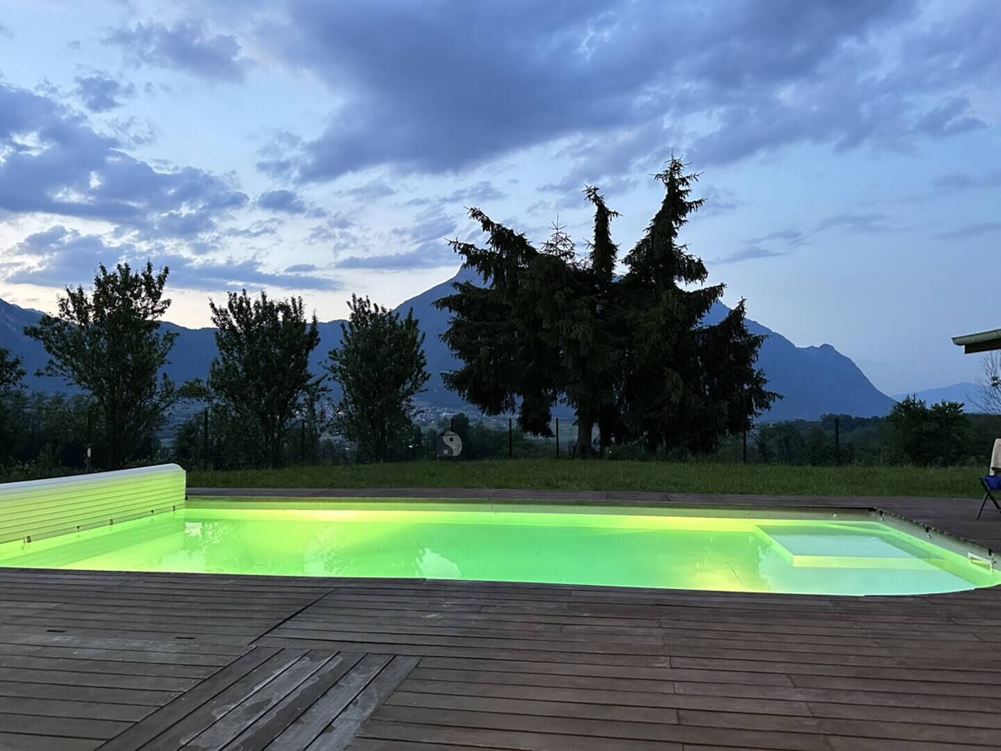 Une piscine avec une terrasse en bois, entourée d’arbres et avec des montagnes en toile de fond.