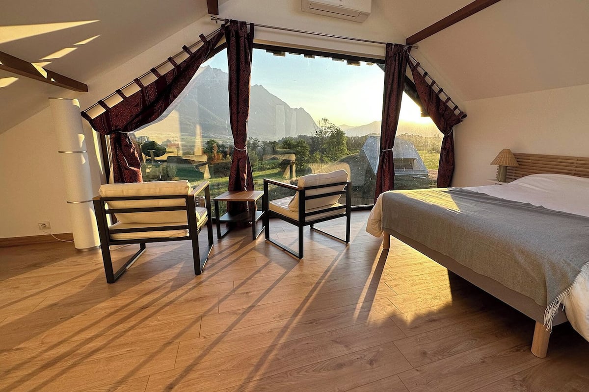 Un dormitorio con cama, dos sillas, una mesita y vistas a montañas a través de amplios ventanales.