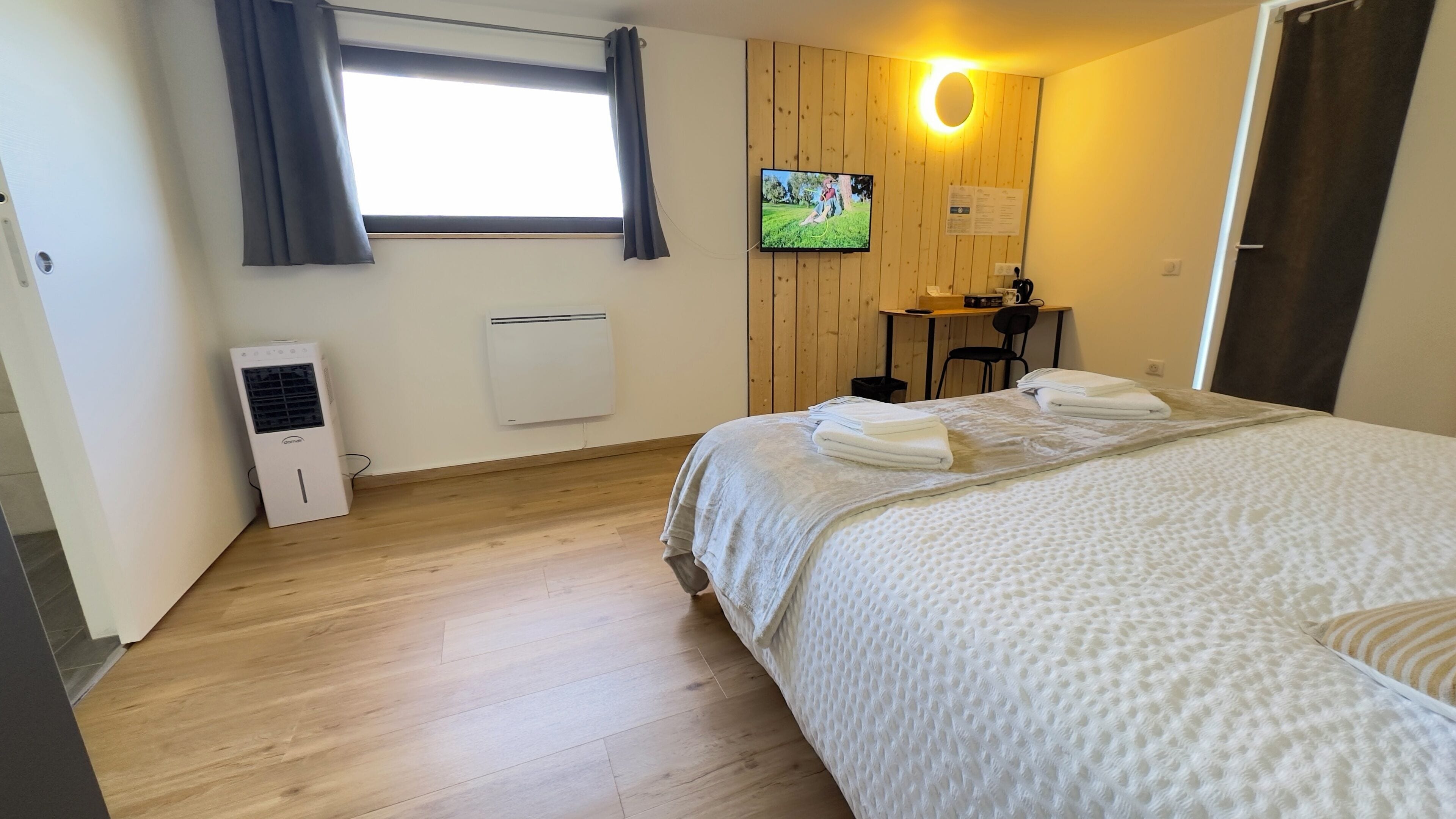 Quarto de hotel com cama, escrivaninha, cadeira, televisão e janela com cortinas.
