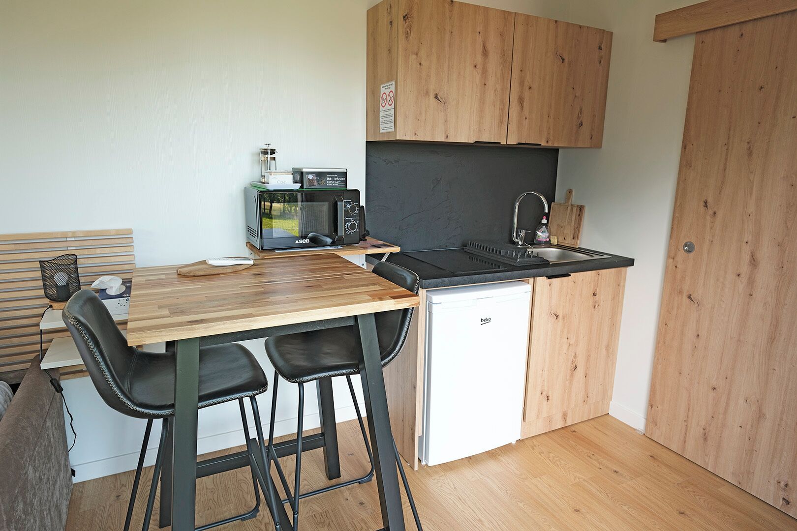 Een compacte kitchenette met een magnetron, spoelbak en kleine koelkast. Twee zwarte barstoelen en een houten tafel zijn aanwezig.
