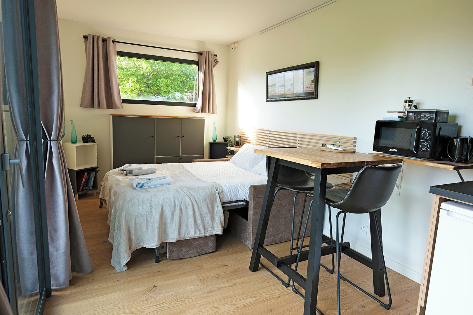 A compact bedroom with a bed, bedside table, and a desk with a chair. A window with curtains and a picture frame are visible.