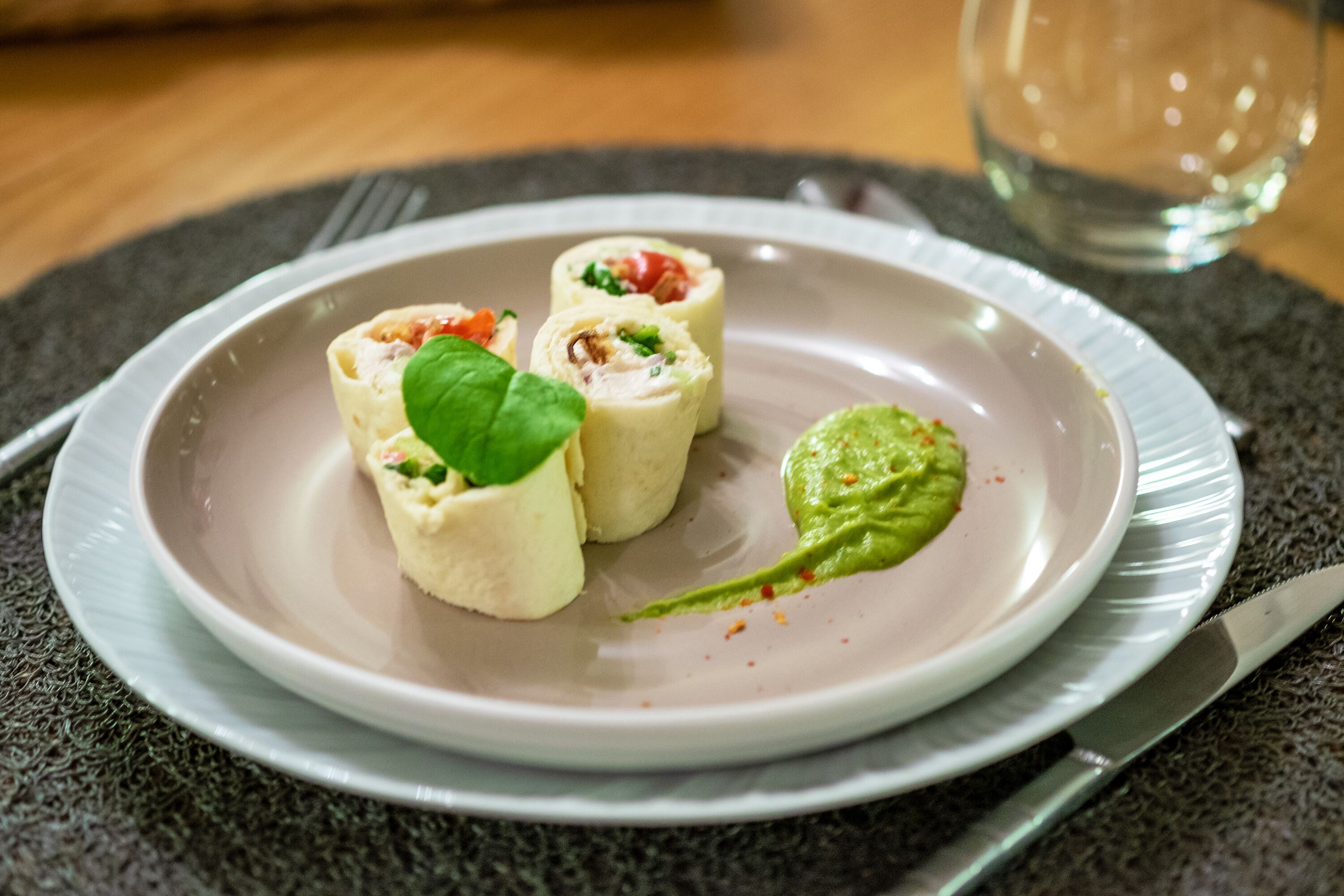 Une assiette contenant des amuse-gueules enroulés, une feuille de basilic et une cuillerée de sauce verte, le tout posé sur un set de table texturé.