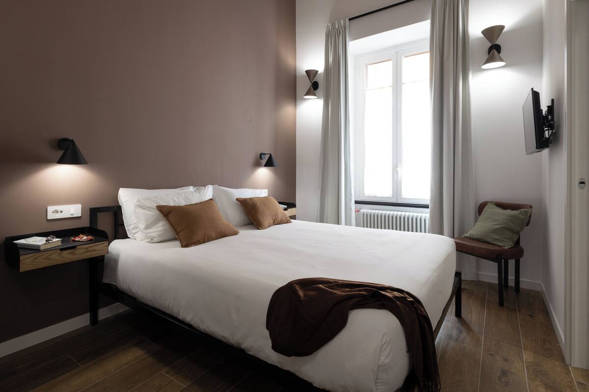 A neatly made bed with white linens and brown pillows, a bedside table with a lamp, and a view of a window with curtains.