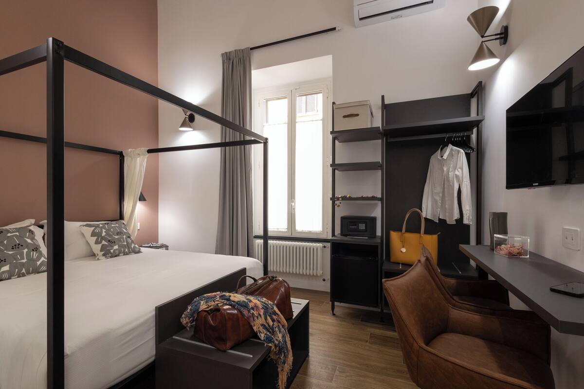 A modern hotel room with a four-poster bed, a desk with a chair, a wardrobe, and a view of the outside.