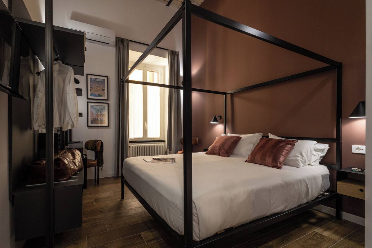 A four-poster bed with a white mattress and pillows, a wooden floor, a desk with a chair, and a coat rack with a jacket.
