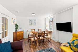 A modern living room with a dining area, a flat-screen TV mounted on the wall, and a wooden cabinet with a plant.