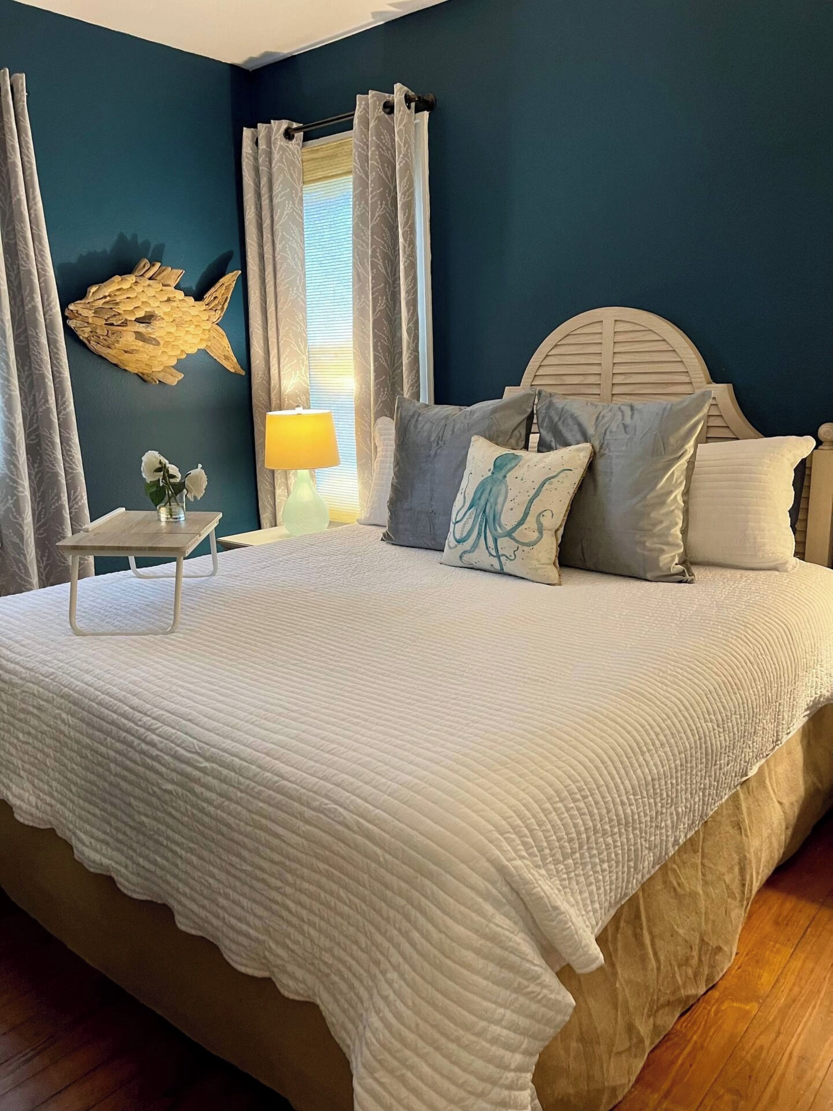 A bedroom with a large bed, a wooden headboard, a wall-mounted fish decoration, a bedside table with a lamp, and a window with curtains.