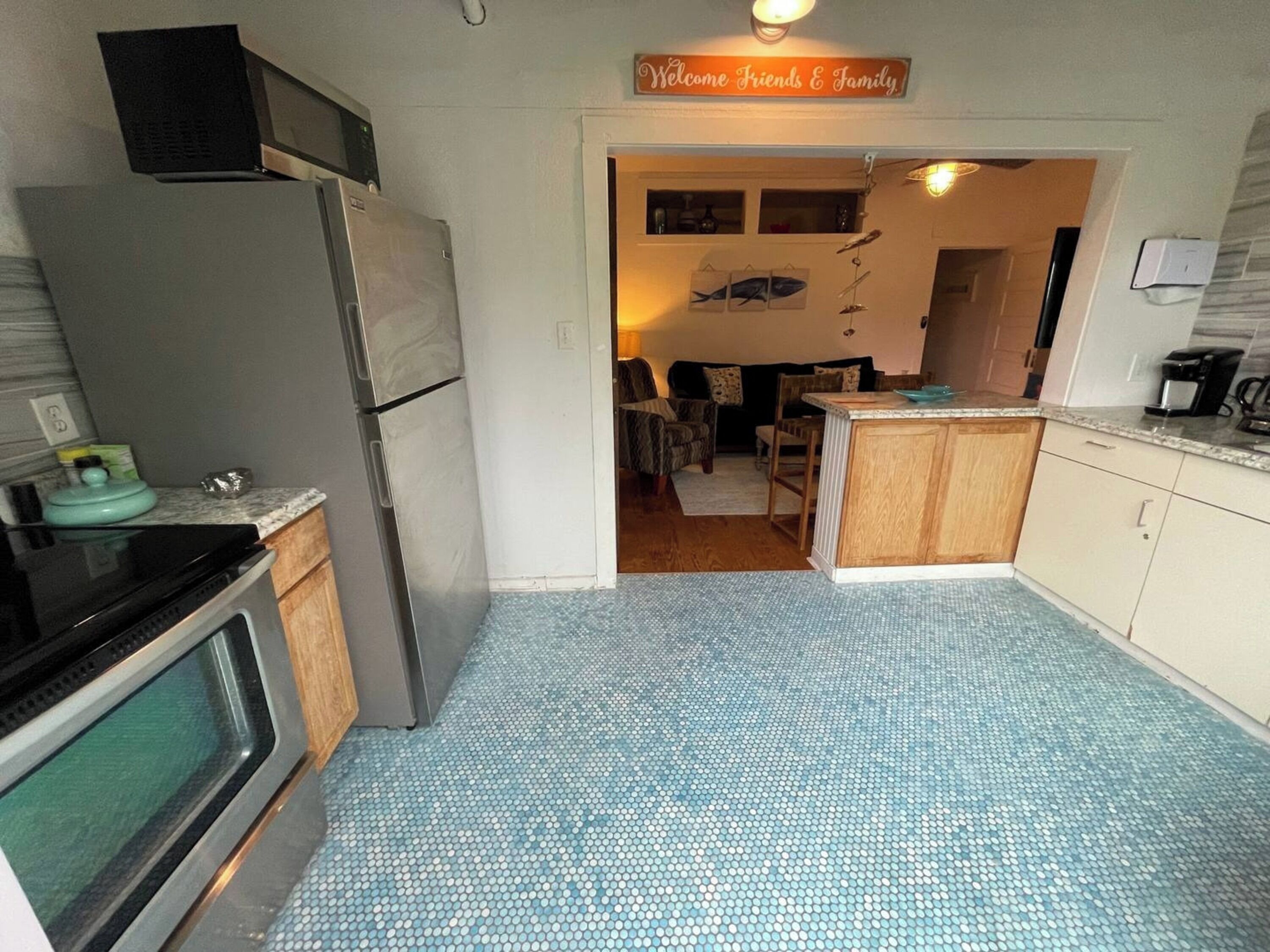 A compact kitchen with a refrigerator, stove, and microwave. A sign above the door reads "Welcome Friends & Family." The floor is tiled, and the kitchen has wooden cabinets.