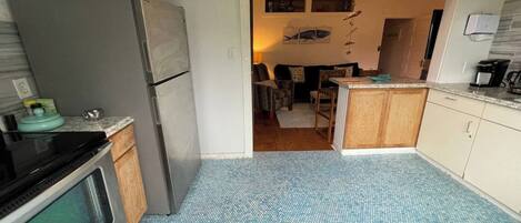A compact kitchen with a refrigerator, stove, and microwave. A sign above the door reads "Welcome Friends & Family." The floor is tiled, and the kitchen has wooden cabinets.