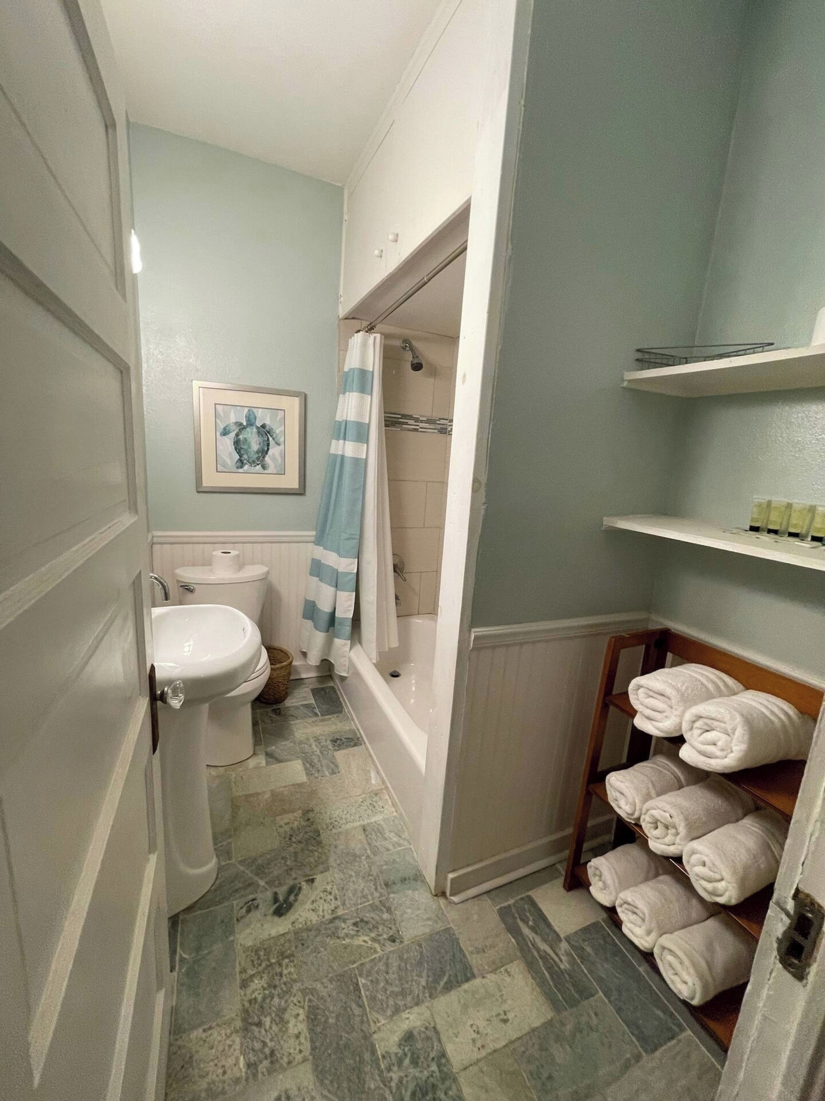 Une salle de bain équipée de toilettes, d’un lavabo, d’une baignoire et d’un rideau de douche. On y trouve des serviettes sur un porte-serviettes et un tableau encadré au mur.
