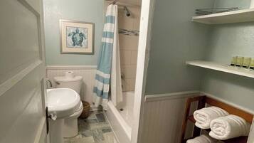 Une salle de bain équipée de toilettes, d’un lavabo, d’une baignoire et d’un rideau de douche. On y trouve des serviettes sur un porte-serviettes et un tableau encadré au mur.