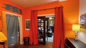 A room with orange walls, a wooden floor, a door with a blue curtain, a small table with a lamp, and a bookshelf.