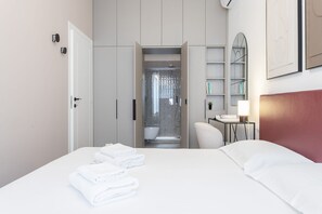 A hotel room with a bed, a bathroom with a shower, a chair, a bedside table with a lamp, and a bookshelf.